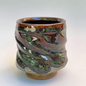 May include: A cylindrical ceramic cup featuring a glossy glaze with swirling patterns of deep brown, forest green, and hints of metallic bronze. The exterior surface has a textured, rippled finish with a smooth, rounded rim and a solid base.