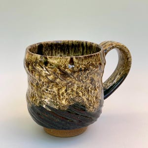 May include: A ceramic mug featuring a textured, rippled surface with a glaze that transitions from a dark, deep brown at the base to a mottled, creamy beige and olive green at the rim. The mug has a sturdy, curved handle and a plain, unglazed base.