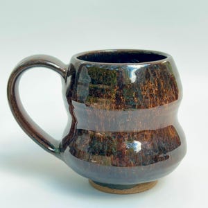 May include: A ceramic mug with a rounded, bulbous body and a single curved handle. The surface features a glossy, dark brown glaze with mottled black and copper-toned streaks throughout, creating a textured, reflective appearance on the exterior.
