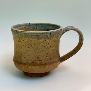 May include: A ceramic mug with a speckled tan and brown body, featuring a flared rim with a blue-grey glaze drip. The mug has a rounded base, a sturdy curved handle on the right side, and a smooth, speckled texture throughout the exterior surface.