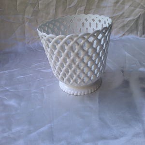 Op de afbeelding: Een witte keramische mand met een rasterontwerp. De mand heeft een ronde basis en een uitlopende bovenkant. Het rasterpatroon creëert een open, luchtig gevoel. De mand wordt waarschijnlijk gebruikt voor decoratieve doeleinden of om kleine items in te bewaren.
