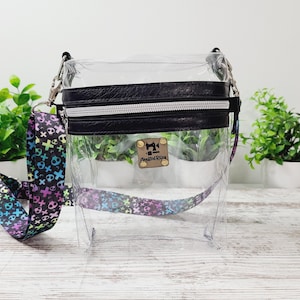 May include: Clear plastic crossbody bag with a black leather flap and zipper. The bag has a rainbow skull and crossbones patterned strap. The bag has a metal tag with the text "Sewing Bliss" on it.