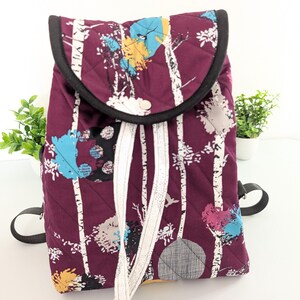 May include: A quilted backpack with a burgundy background and a white birch tree pattern with colorful accents. The backpack has a black trim, black straps, and white drawstring closure. The design includes blue, yellow, and pink details.