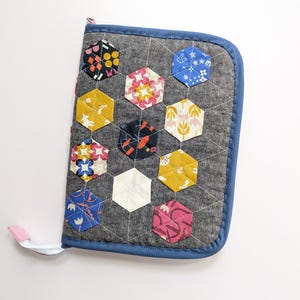 May include: A quilted notebook cover with a gray background and a blue border. The cover features a pattern of colorful hexagonal patches with various designs, including floral and geometric patterns. A pink loop is attached to the side.