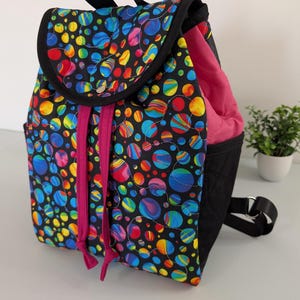 May include: A colorful backpack with a black base and flap. The flap and front panels feature a vibrant pattern of multicolored circles on a black background. The interior is pink, and the bag has black straps and a drawstring closure.