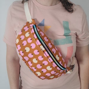 May include: A brown fanny pack with a zipper closure and a white strap. The fanny pack has a pink and white apple print.