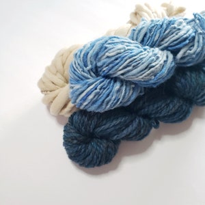 Pode incluir: Close-up de quatro novelos de fio em tons de azul e creme. O fio é espesso e texturizado, com uma aparência grossa. As cores variam de um azul marinho profundo a um creme claro, criando um efeito de gradiente. O fio é provavelmente para tricô ou crochê.