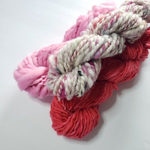 Puede incluir: Tres ovillos de hilo en tonos de rosa, rojo y blanco. El hilo está dispuesto en una pila, con el hilo blanco en la parte superior.