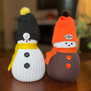 May include: Two sock snowmen with team logos. One snowman is wearing a black hat with a Pittsburgh Steelers logo and a yellow scarf. The other snowman is wearing an orange hat with a Cleveland Browns logo and an orange scarf.