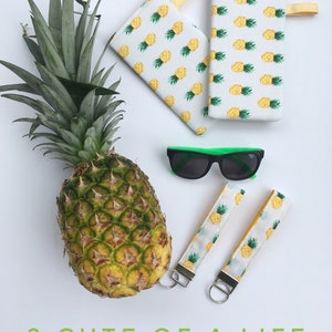 Könnte beinhalten: Zwei Schlüsselanhänger mit Ananas-Muster, gelbem Besatz und silbernen Schlüsselringen. Ein Brillenetui mit Ananas-Muster und gelber Klappe. Eine grüne Sonnenbrille. Eine ganze Ananas auf weißem Hintergrund. Der Text "2 CUTE OF A LIFE" ist in Grün unter den Artikeln gedruckt.
