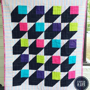 May include: A black and white quilt with a geometric pattern of squares in pink, green, purple, and blue. The quilt is edged with a pink and white striped fabric. The text "2 CUTE OF A LIFE" is in a circle in the lower right corner.