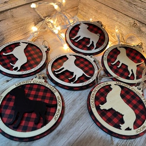 May include: Six round wooden ornaments with red and black buffalo plaid designs. Each ornament features a different dog breed silhouette and the breed name written around the edge. The text "Merry Christmas Woof Woof" is written on each ornament.