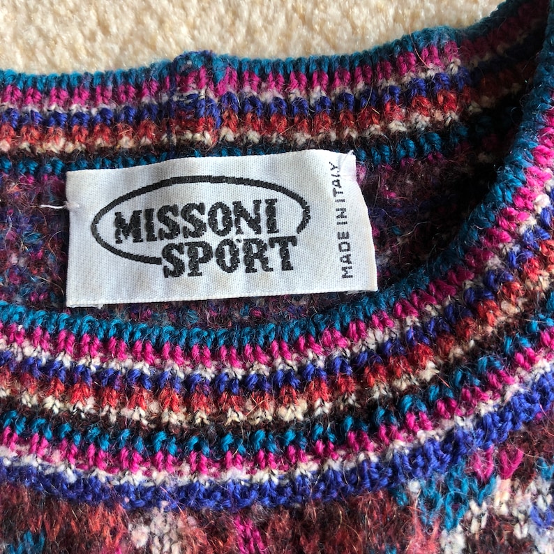 May include: A close-up of a colorful knitted sweater with a Missoni Sport label. The label is white with black text and a black oval around the word "Missoni". The label also says "Made in Italy".