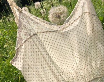Puede incluir: Un vestido blanco de encaje con un patrón de lunares y un borde de encaje. El vestido está colgado en un campo de hierba verde y flores de diente de león blancas.