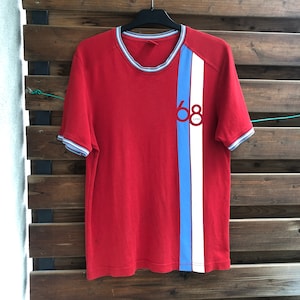 May include: Red t-shirt with a vertical stripe in light blue and white. The shirt has a crew neck and short sleeves with blue and white trim. The number "68" and the word "ESPRIT" are printed on the front.