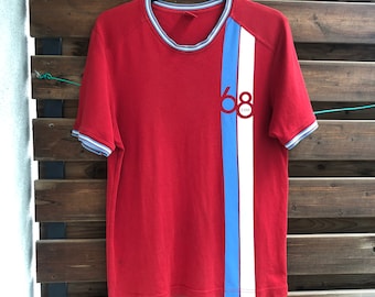 May include: Red t-shirt with a vertical stripe in light blue and white. The shirt has a crew neck and short sleeves with blue and white trim. The number "68" and the word "ESPRIT" are printed on the front.