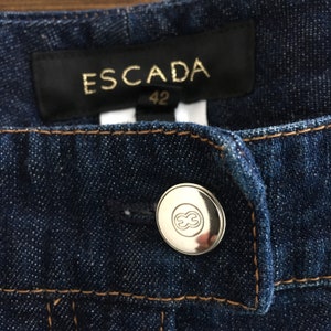 May include: Close-up of a dark blue denim fabric with a brown thread seam. A silver button with the Escada logo is visible. The label reads "ESCADA 42".