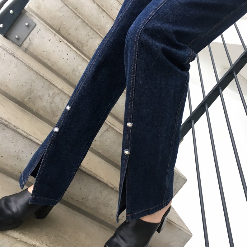 May include: Dark blue denim jeans with a slit on each leg and silver button accents.