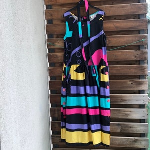 Pu&ograve; includere: Un abito vintage senza maniche, con sfondo nero e forme geometriche colorate. L'abito presenta una gonna a righe con bande orizzontali gialle, viola, turchesi e nere. I bottoni rosa adornano il lato e una cravatta abbinata pende dallo scollo.