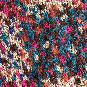 May include: Close-up of a knitted fabric with a colorful geometric pattern. The fabric is made of wool and has a soft, fuzzy texture. The colors in the pattern are brown, blue, pink, and white.
