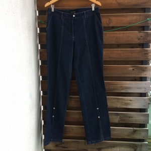 May include: Dark blue denim jeans with a straight leg and decorative buttons on the sides. The jeans have a brand label that says "ESCADA" on the back waistband.