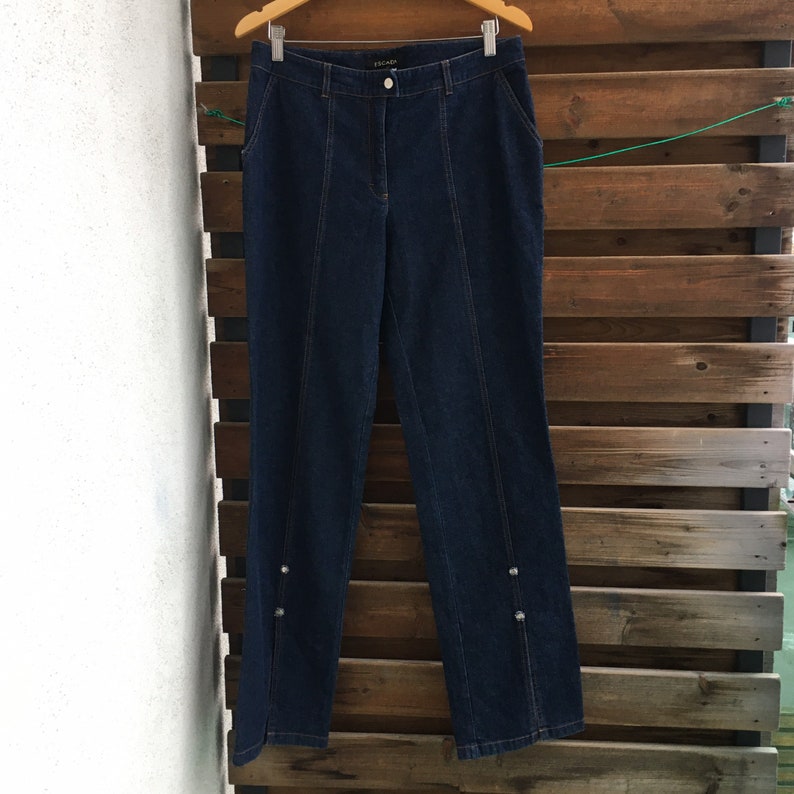 May include: Dark blue denim jeans with a straight leg and decorative buttons on the sides. The jeans have a brand label that says "ESCADA" on the back waistband.