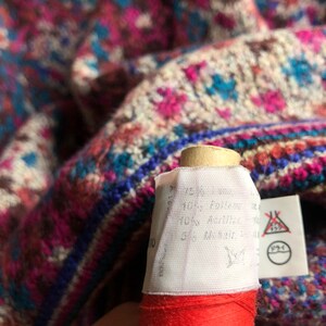 May include: Close-up of a white fabric label with black text. The label is attached to a red and white spool of thread. The text on the label reads "Made in Germany, 100% Polyester, 40&deg;C Machine Wash, 5% Acrylic, Do Not Bleach, Do Not Tumble Dry, Iron at Low Temperature, Do Not Dry Clean".