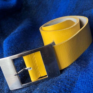 May include: A yellow leather belt with a silver buckle. The belt is rolled up and the buckle is on the side.