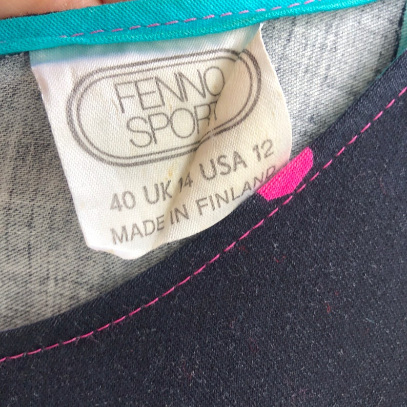 Pu&ograve; includere: Primo piano di un'etichetta di abbigliamento con il marchio "FENNO SPORT". L'etichetta indica la taglia 40 (Europa), UK 14 e USA 12. Il capo &egrave; prodotto in Finlandia. Il capo &egrave; blu navy con cuciture rosa.