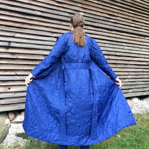 May include: A long, blue quilted coat with a floral pattern. The coat has a belt and a long, flowing design.