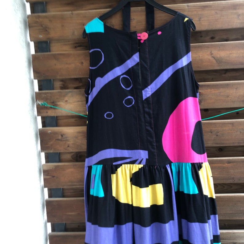 Pu&ograve; includere: Abito nero senza maniche con un motivo geometrico colorato. L'abito presenta un design vibrante con forme rosa, viola, gialle e turchese. Ha una chiusura con bottoni e un orlo a balze, appeso a una gruccia nera.