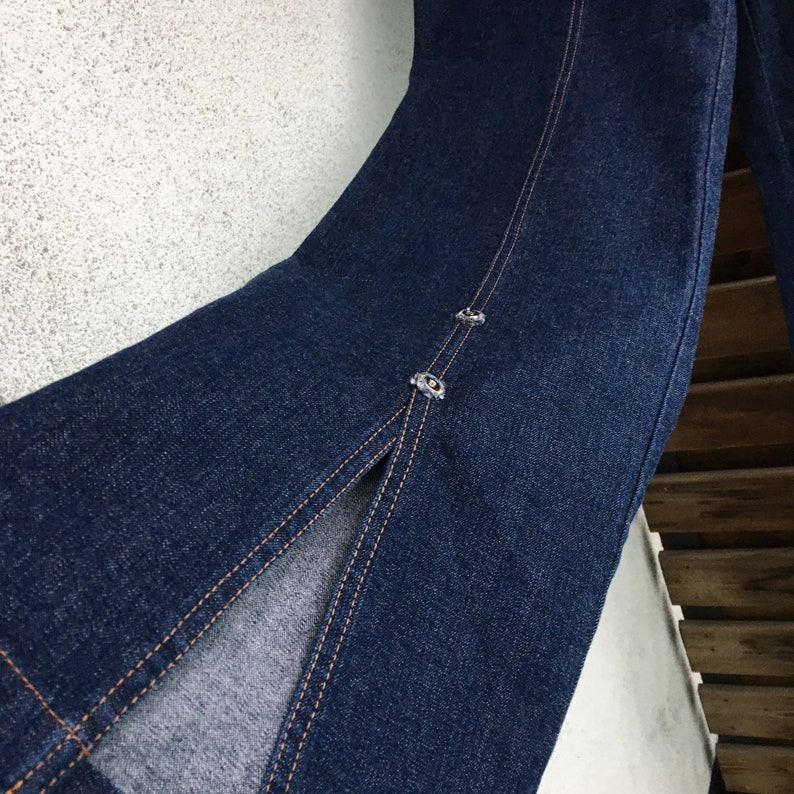 May include: A pair of dark blue denim jeans with a long slit up the front of each leg. The jeans have a seam down the center of the front and two small metal buttons on each leg.