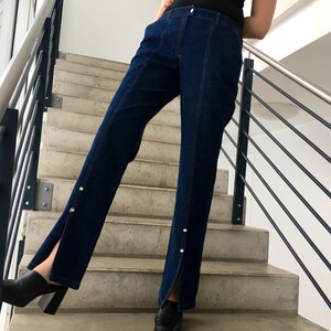 May include: A pair of dark blue denim jeans with a slit up the side of each leg. The jeans have a high waist and are fitted through the hips and thighs. The jeans are worn by a person standing on a set of stairs.