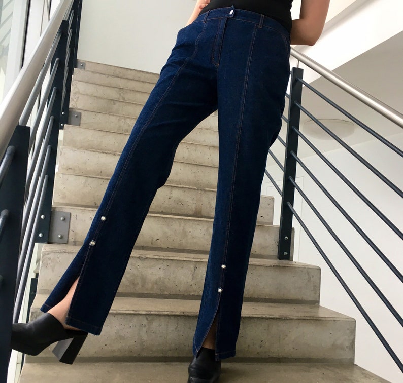 May include: A pair of dark blue denim jeans with a slit up the side of each leg. The jeans have a high waist and are fitted through the hips and thighs. The jeans are worn by a person standing on a set of stairs.