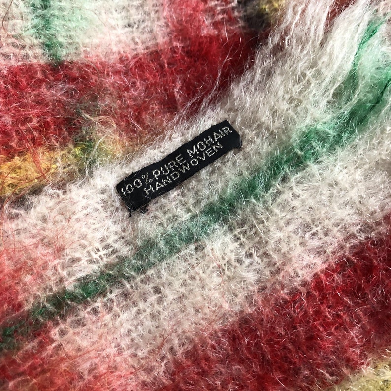 May include: Close-up of a multi-coloured, fuzzy, striped scarf with a black label that reads "100% Pure Mohair Handwoven".