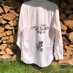 Può includere: Una camicia bianca con bottoni e strisce rosa con una stampa di quattro auto d'epoca e un trattore sul retro.