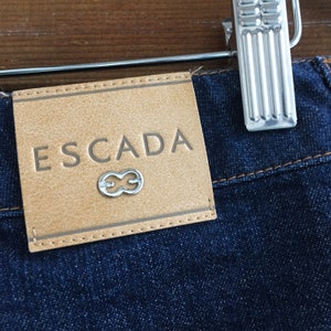 May include: A brown leather patch with the word "ESCADA" and a silver infinity symbol on a dark blue denim background.