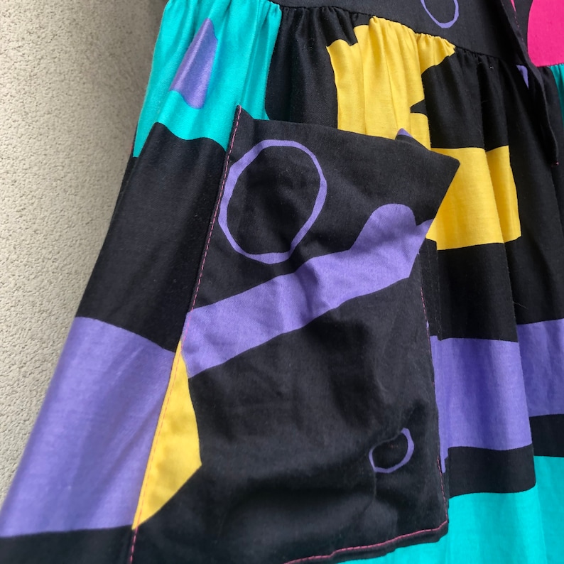 Pu&ograve; includere: Una gonna colorata con un motivo geometrico. La gonna presenta grandi blocchi di nero, turchese, giallo e viola. Una tasca con un motivo a cerchio viola &egrave; visibile sul davanti. La gonna &egrave; realizzata in tessuto leggero.