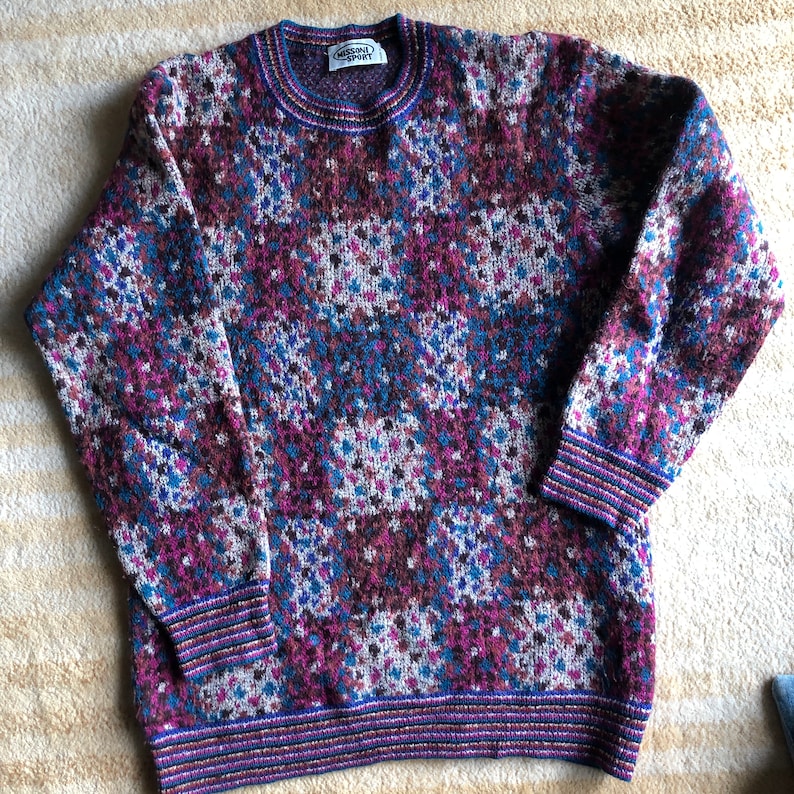 May include: A multi-colored sweater with a geometric pattern in shades of purple, blue, brown, and white. The sweater has a crew neck and ribbed cuffs and hem. The label on the sweater reads "Worsted Sport".