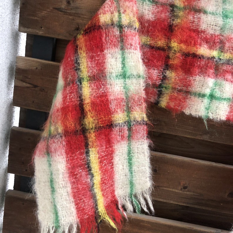 May include: A red, white, green and yellow plaid scarf with a fuzzy texture. The scarf is folded and draped over a wooden surface.