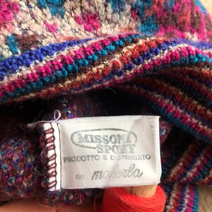 May include: Close-up of a white tag with black text that reads "Missoni Sport" and "Prodotto e distribuito da Manifattura". The tag is attached to a colorful knitted fabric with stripes of pink, blue, brown, and white.
