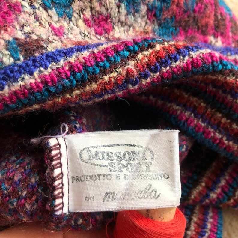 May include: Close-up of a white tag with black text that reads "Missoni Sport" and "Prodotto e distribuito da Manifattura". The tag is attached to a colorful knitted fabric with stripes of pink, blue, brown, and white.
