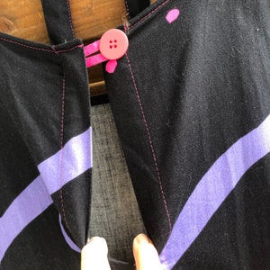 Pu&ograve; includere: Primo piano di un capo in tessuto nero con strisce diagonali lavanda e cuciture rosa. Un bottone rosa chiude l'apertura del capo. Le cinghie sono nere. Il capo sembra fatto a mano.