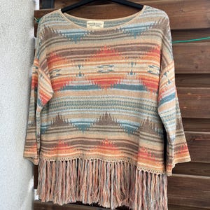 May include: A brown, orange and blue striped sweater with a fringe trim. The sweater has a crew neck and long sleeves. The brand label reads "South & Supply".