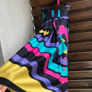 Pu&ograve; includere: Abito senza maniche con un vivace motivo astratto in nero, giallo, viola, turchese e rosa. L'abito ha una gonna fluida, un fiocco al collo e bottoni rosa. L'abito &egrave; appeso a una gruccia nera.