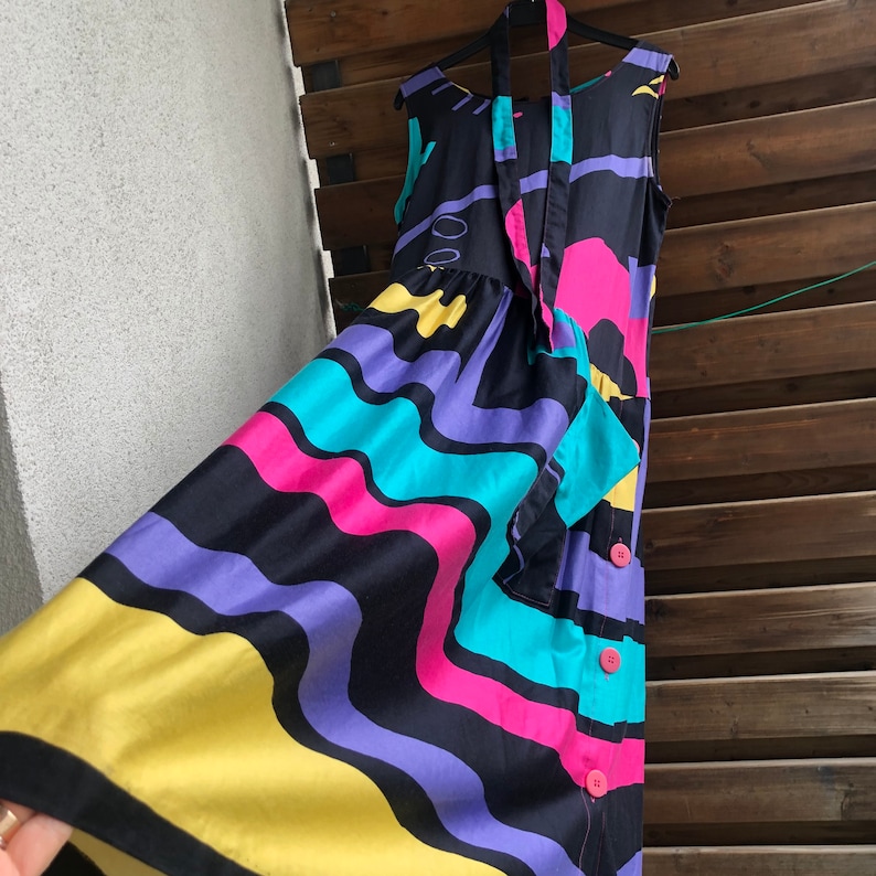 Pu&ograve; includere: Abito senza maniche con un vivace motivo astratto in nero, giallo, viola, turchese e rosa. L'abito ha una gonna fluida, un fiocco al collo e bottoni rosa. L'abito &egrave; appeso a una gruccia nera.