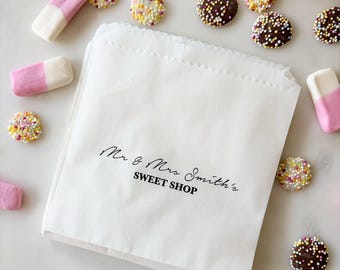 May include: A white paper bag with the text "Mr & Mrs Smith's SWEET SHOP" in black script. Scattered around the bag are pink and white marshmallow candies and chocolate candies covered in colorful sprinkles.