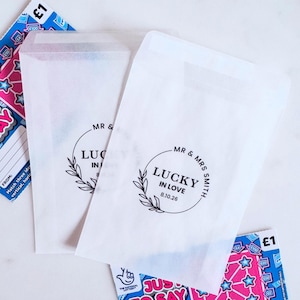 May include: Two white paper bags with the text "MR & MRS SMITH LUCKY IN LOVE 8.10.26" in a circular design. The bags are surrounded by lottery tickets with blue, pink, and white designs. The bags are likely for a wedding or special event.