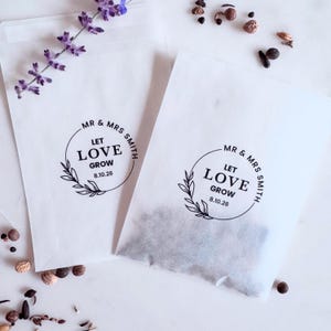 Op de afbeelding: Twee witte papieren zaadzakjes met de tekst "MR & MRS SMITH LET LOVE GROW 8.10.26" in een cirkelvormig ontwerp. Eén zakje is gevuld met zaden. Een lavendeltakje en losse zaden zijn verspreid.