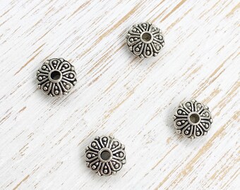 May include: Four silver metal spacer beads with a decorative floral design. The beads have a round shape with a hole in the center.